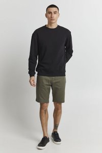 Sweatshirt noir à col rond, associé à un short vert olive. Le modèle porte des baskets noires et a des tatouages minimalistes sur le bras visible.