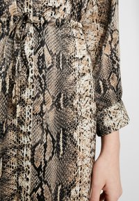 Långärmad klänning i ormskinnsmönster med nyanser av beige, brunt och svart. Har en knuten midja och manschetterade ärmar. Slät textur.