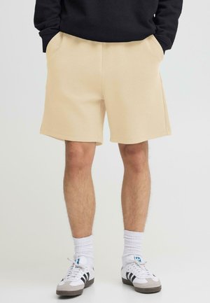 Mand iført beige shorts, hvide crew-sokker, hvide sneakers med sorte striber og en sort sweatshirt med hænderne i lommerne.