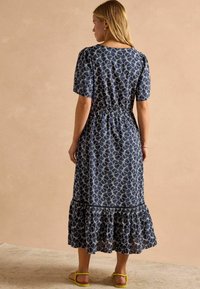 Marineblaues Kleid mit floralem Muster, kurzen Puffärmeln, tailliertem Schnitt und gestuftem Rüschensaum. Getragen mit gelben Sandalen.