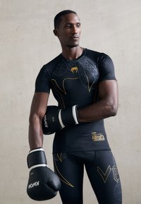 Sort atletisk skjorte med gule grafiske accenter, parret med sorte boksehansker og matchende leggings, der har et struktureret design.
