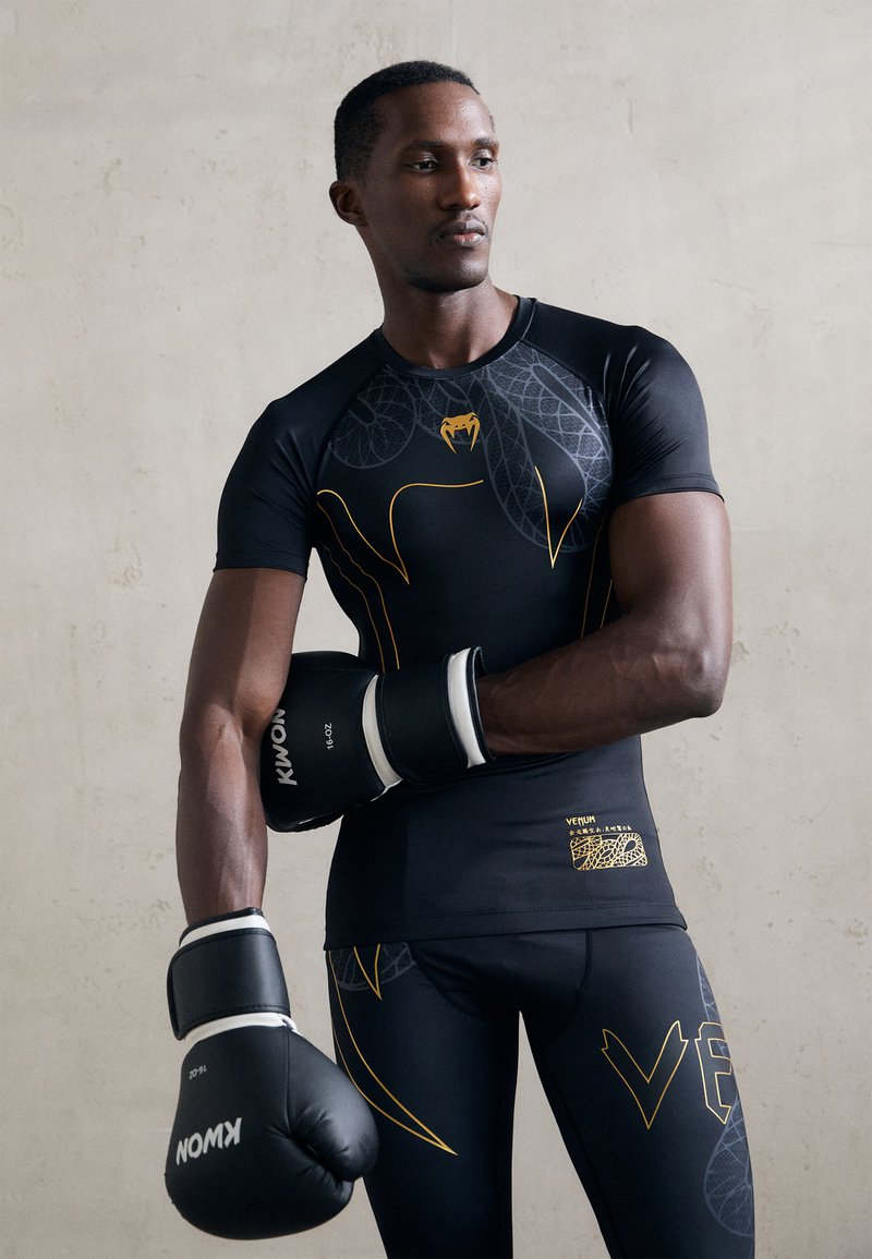 Sort atletisk skjorte med gule grafiske accenter, parret med sorte boksehansker og matchende leggings, der har et struktureret design.
