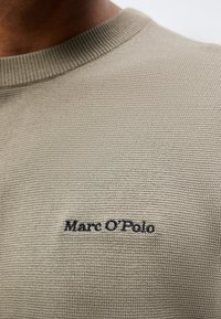 Κοντινό πλάνο μπεζ πλεκτού πουλόβερ με ριμπ καβάλο και μαύρο κεντητό λογότυπο "Marc O'Polo" στο στήθος.