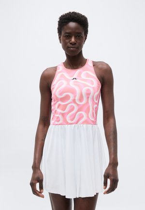 Woman wearing sleeveless pink top with white abstract patterns and a white pleated skirt, standing against a plain light background.