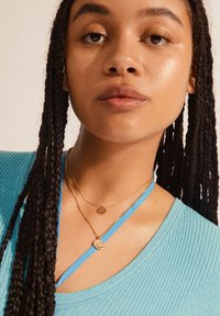 Teal ribbed top, layered gold necklaces, and blue ribbon accent. Skin appears moisturized; braided hair adds texture to the overall look.