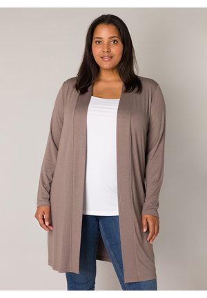 Vrouw met medium-donkere huid en donker haar draagt een taupekleurige lange cardigan over een wit topje en blauwe jeans, staand tegen een lichte achtergrond.