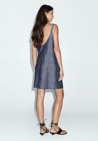 Vestito di jeans, colore blu scuro, senza maniche con un design a schiena bassa, lunghezza al ginocchio, realizzato in tessuto leggero, abbinato a sandali neri con cinturini.