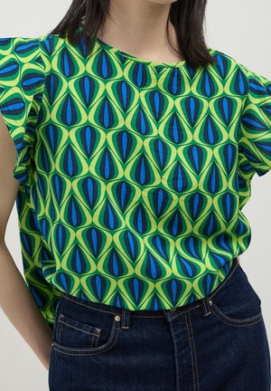 Vrouw draagt een groen T-shirt met korte mouwen met een blauw en donkergroen geometrisch patroon, in een donkerblauwe spijkerbroek gestopt.