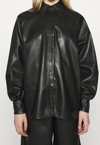 Camicia nera in pelle con bottoni, colletto a bande, maniche raglan e polsini arricciati. Tessuto liscio con una finitura lucida.