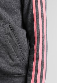 Sweat-shirt gris avec un design à trois bandes roses sur la manche, doté d'une poche latérale. Texture douce et coupe décontractée.