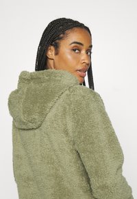 Flauschige, olivegrüne Kapuzenjacke mit strukturiertem Finish, die einen lässigen Schnitt und eine ausgeprägte Kapuze aufweist.