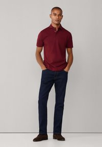 Bordová polosvetrová košeľa s krátkymi rukávmi, skombinovaná s tmavomodrými denimovými džínmi a hnedými semišovými topánkami. Model stojí s rukami v vreckách.