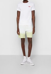 T-shirt blanc à manches courtes avec un logo de marque rouge sur la poitrine, associé à un short dégradé vert. Porté avec des baskets montantes blanches.