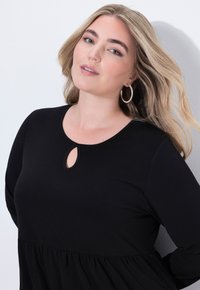 Kobieta z długimi blond włosami, nosząca duże kolczyki w kształcie koła i czarną bluzkę z małym dekoltem w kształcie łezki, pozująca na tle białej ściany.