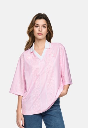 Frau mit lockerem welligem Haar, die ein hellrosa gemustertes kurzärmliges Poloshirt mit weißem Kragen und blaue Jeans trägt, Hand in der Tasche.
