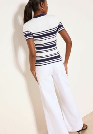 Žena s tamnim repom nosi kratku majicu s mornarsko plavim i bijelim prugama, široke bijele hlače i crne sandale na petu, stoji naslonjena na svijetli zid.