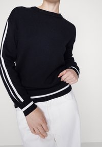 Pull en maille bleu marine avec des rayures blanches le long des manches et des poignets et ourlets côtelés, associé à un pantalon blanc.