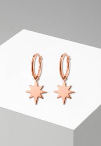 Boucles d'oreilles créoles en or rose avec des pendentifs en forme d'étoile. Le design présente une finition lisse et des lignes épurées, avec une texture polie.