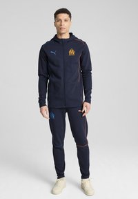 Tuta navale con felpa con zip e pantaloni. Presenta applicazioni arancioni e azzurro chiaro, logo sul petto e polsini elasticizzati. Indossata con sneakers beige.