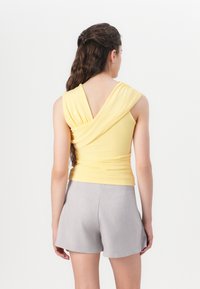 Top sans manches jaune avec un dos drapé, présentant un tissu froncé. Associé à un short gris clair, les deux en tissu lisse et texturé.
