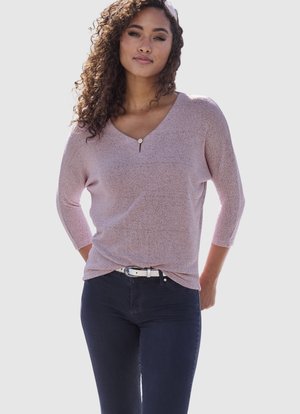 Frau mit lockigem Haar, die ein hellrosa V-Ausschnitt-Oberteil mit 3/4-Ärmeln, dunkle Hosen und einen weißen Gürtel trägt, stehend mit den Händen hinter dem Rücken.