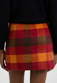 Mini gonna in lana con un motivo a quadri rosso, arancione e marrone. Presenta una forma aderente e orlo piatto, con pieghe visibili sul retro.