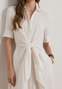 Fehér lenvászon ingruha rövid ujjakkal, elöl megkötős részlettel és gombos gallérral. Simább textúrájú, laza szabású.