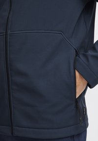 Marineblå softshell-jakke med glidelommer, teksturert stoff og en tettsittende design. Har glidelås åpen og merket tag nær kanten.