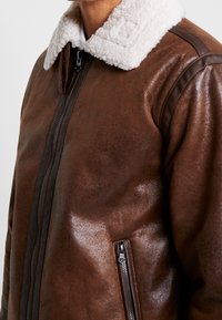 Bruine leren bomberjack met een witte shearling kraag, ritssluiting, zijritszak en een gestructureerde afwerking. Voorzien van donkerbruine schouderaccenten.