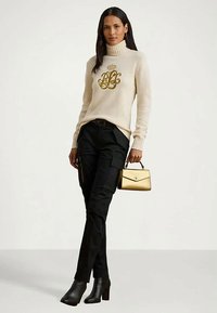 Κρεμ ριμπάριστο πουλόβερ με χρυσό μονογράμμα, μαύρο cargo παντελόνι και χρυσή τσάντα. Το μοντέλο φορά μαύρες μποτάκια μέχρι τον αστράγαλο, στέκεται σε ουδέτερο φόντο.