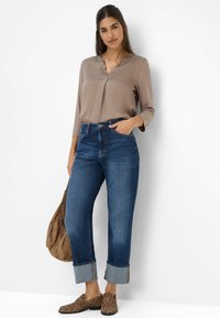 Frau in einer taupefarbenen Bluse, mit hochgekrempelten blauen Jeans und Loafers im Leopardenmuster, die eine braune Handtasche hält und vor einer weißen Wand steht.