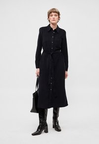 Černé košilové šaty z texturované látky, s dlouhými rukávy, páskem v pase a náprsní kapsou. Kombinované s kozačkami ke kolenům.