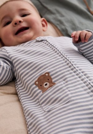 Bambino sorridente sdraiato che indossa una tutina a righe grigie e bianche con un piccolo orsetto marrone sulla tasca frontale.