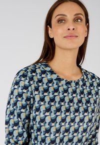 Blauw patroon top met geometrische ontwerpen in teal, marineblauw en goud. Ronde halslijn en textuurstof.