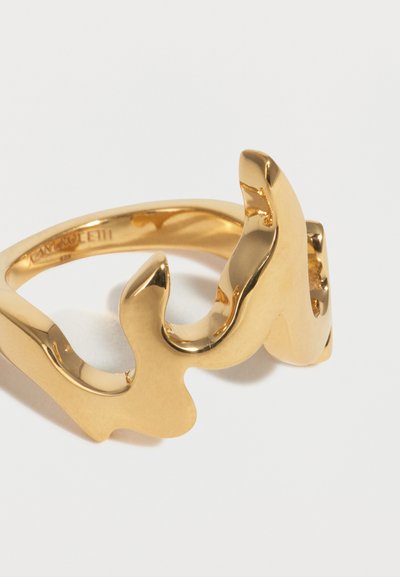 Δαχτυλίδι από μέταλλο με χρυσή απόχρωση, με αφηρημένο, κυματιστό σχέδιο σε σχήμα φλόγας σε απλό λευκό φόντο.