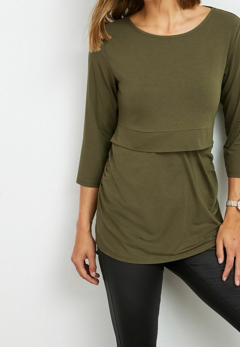 Olivgrünes Langarm-Oberteil mit runder Halslinie und geschichtetem Frontdesign, gefertigt aus weichem, dehnbarem Stoff. Tragen mit schwarzen Leggings.
