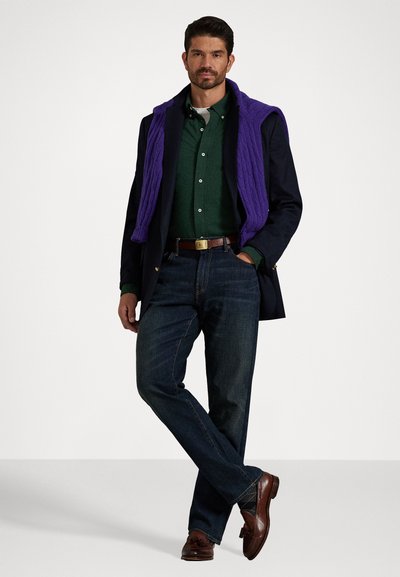Mann iført mørk blazer, grønn skjorte, mørke jeans, brun belte og sko, med lilla genser drapert over skuldrene, stående med bena krysset.