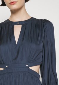 Gros plan d'une personne portant une robe bleu foncé à manches longues avec un décolleté troué et des découpes latérales maintenues par de petits boutons métalliques.