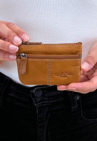 Braune Lederkartenbörse mit Reißverschluss, Vordertasche und geprägt Logo. Die Nähte sind sichtbar. Kompakte, rechteckige Form.
