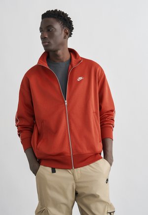 Tânăr cu păr creț și închis la culoare, purtând o jachetă roșie Nike cu fermoar și pantaloni cargo bej, stând cu mâinile în buzunare, privind spre stânga.