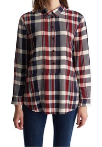 Donna che indossa una camicia a quadri con maniche lunghe e bottoni nei colori blu navale, rosso e crema, abbinata a jeans blu scuro, in piedi contro uno sfondo bianco.