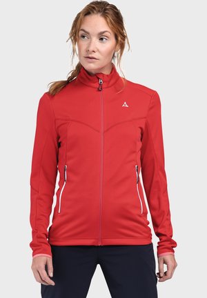 Woman with red zip-up jacket and black pants looking slightly to her left against a plain light gray background.