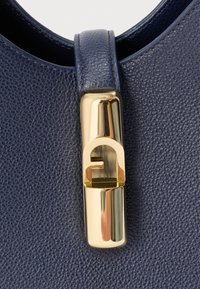 Jūras zilas krāsas ādas rokassomiņa ar teksturētu virsmu, kurai ir zelta tonīša aizdare ar raksturīgu logotipa dizainu un gludiem akcentiem.
