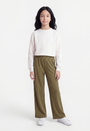 Meisje met lang zwart haar draagt een witte trui, olijfgroene wijde broek en witte sneakers, staand tegen een effen witte achtergrond.