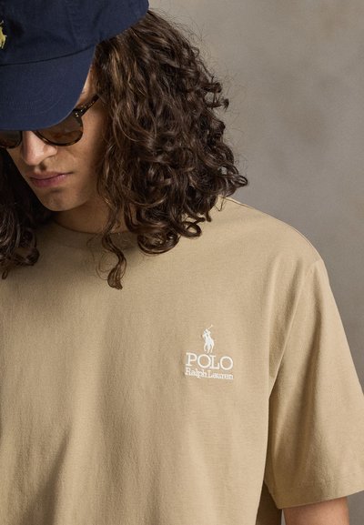 Νέος με κατσαρά μαλλιά που φοράει φιμέ γυαλιά, σκουρόχρωμο καπέλο και μπεζ T-shirt Polo Ralph Lauren με λογότυπο στο στήθος.