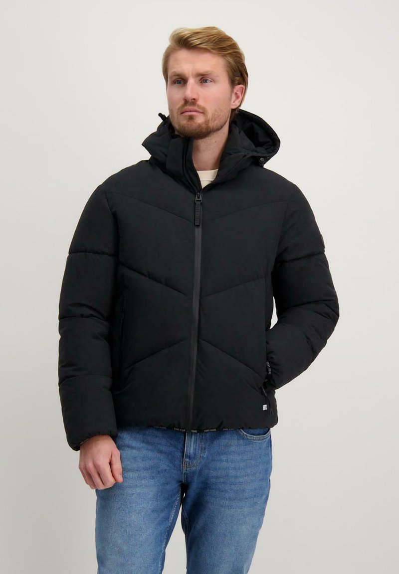 Zwarte gewatteerde jas met een capuchon, voorzien van een diagonaal chevronpatroon, een rits aan de voorkant en elastische manchetten. Draagt met blauwe jeans.
