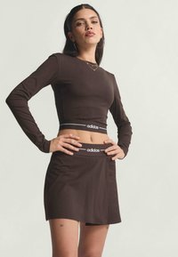 Conjunto de top corto de manga larga y falda en marrón, hecho de una tela suave. Presenta el logotipo blanco de Adidas en la cinturilla. Ajuste ceñido al cuerpo, con costuras mínimas.