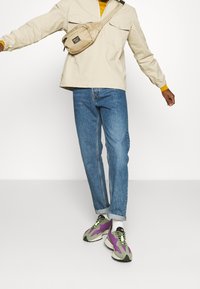 Chaqueta beige con cremallera, interior amarillo, vaqueros azul claro y zapatillas multicolor con detalles en púrpura, verde y negro. Bolso cruzado en beige.