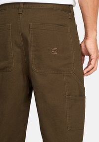 Bruine cargobroek gemaakt van duurzaam materiaal, met meerdere zakken, een soepel textuur en een geborduurd logo op de achterzak.