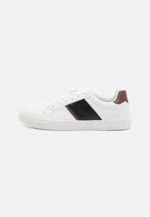 Białe sneakersy z teksturowaną gumową podeszwa, z czarną ukośną linią oraz brązowym skórzanym akcentem na pięcie. Zaprojektowane do codziennego noszenia.